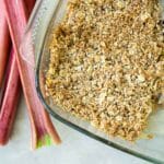 Overhead image of vegan Rhubarb & Apple Crumble. image shows crumble in a glass dish with a silver cake slice, surround by some stalks of rhubarb.