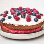 Vegan Gluten Free Sponge Cake. Cake is on a light grey plate on a white background and is decorated with raspberry jam, coconut whipped cream, blueberries and raspberries.