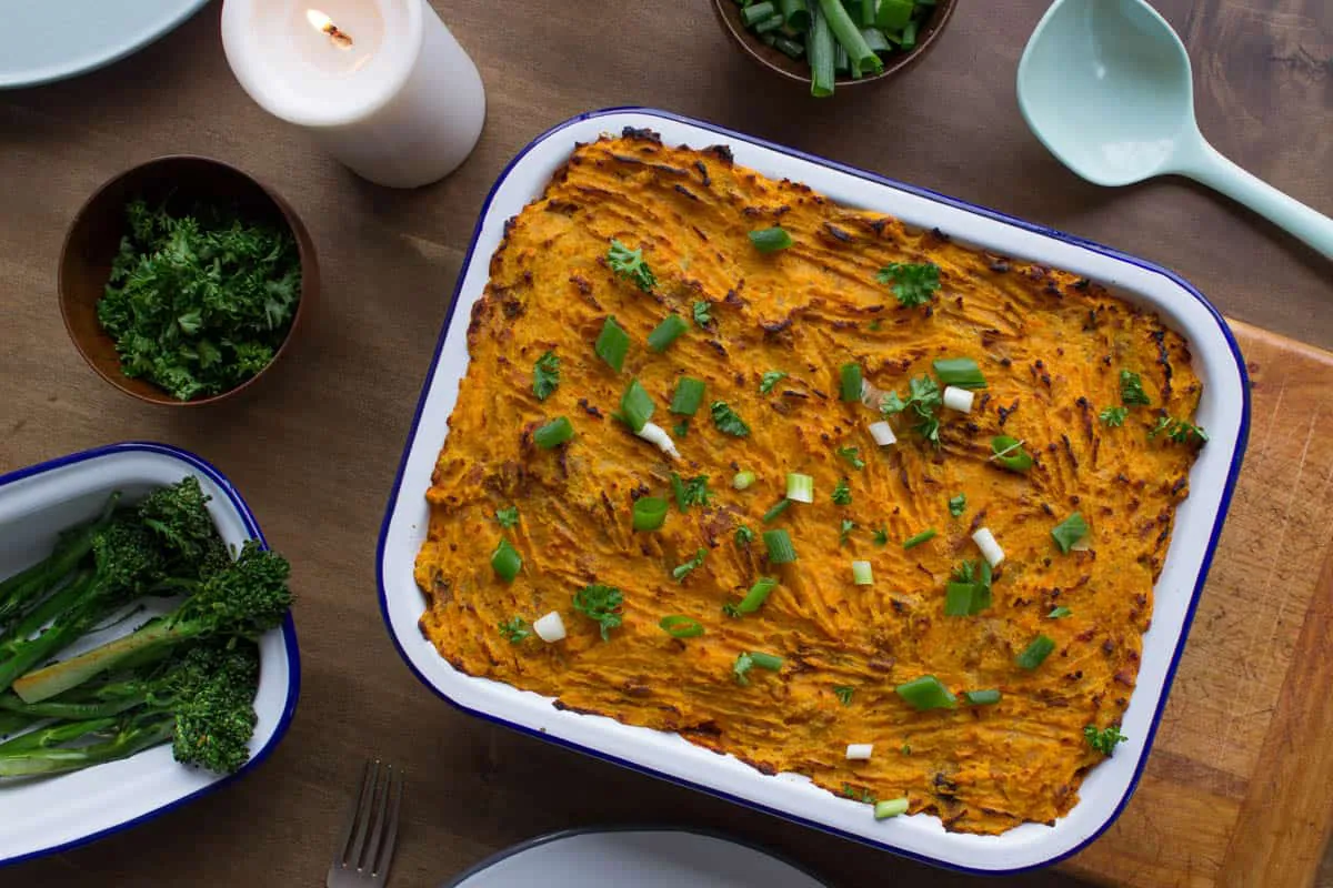 Overhead shot of Vegan Shepherd's Pie with Yam Mash, on chopping board surrounded by serving spoon, bowls and candles.