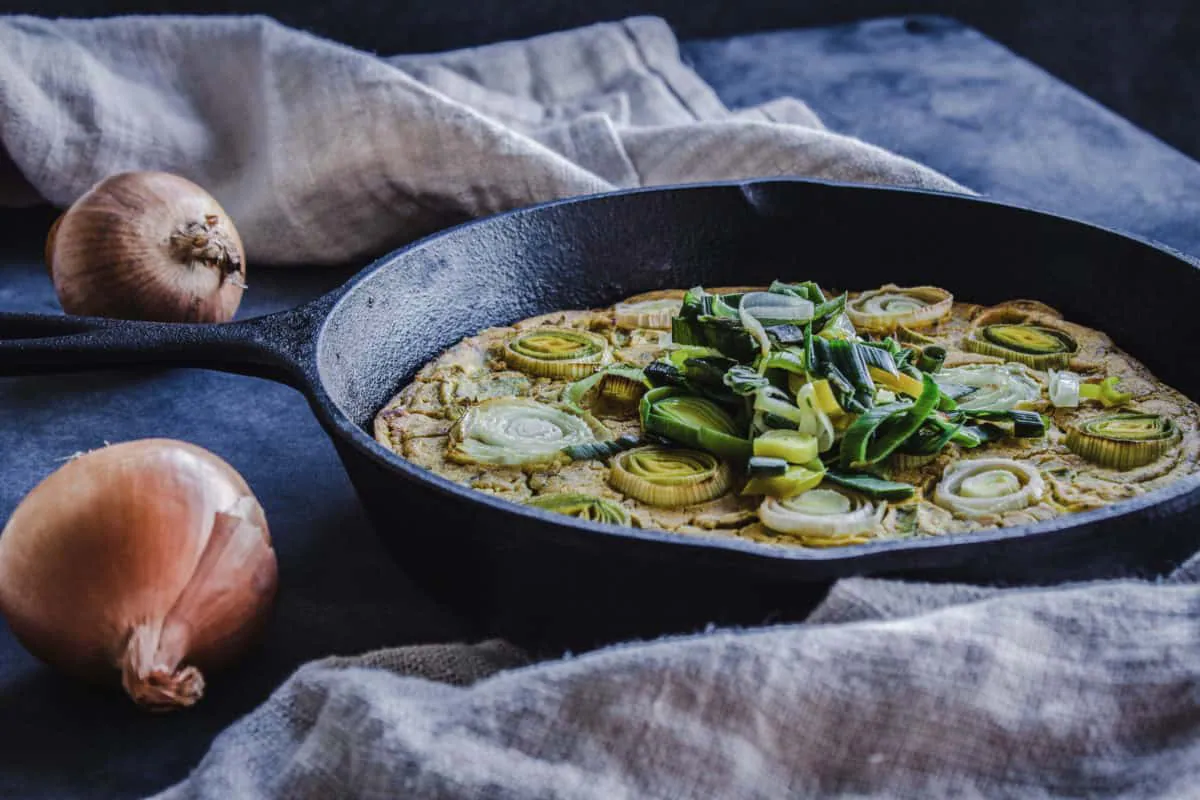 Side shot of Vegan Leek & Onion Frittata in cast iron skillet. Frittata is decorated with leek and onion discs, and is surrounded by onions and linen napkins.