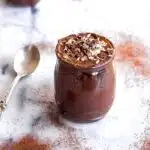 Image of Avocado Chocolate Mousse (vegan & gf). Image shows one pot of chocolate mousse on a white marble table, surrounded by piles of cacao powder and an antique silver spoon.