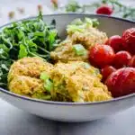 Easy Vegan Chickpea Fritter Bowls on a white marble background. Bowl is filled with chickpea fritters, roasted cherry tomatoes, arugula and hummus. Tomatoes, chickpeas and arugula are scattered around the bowl.
