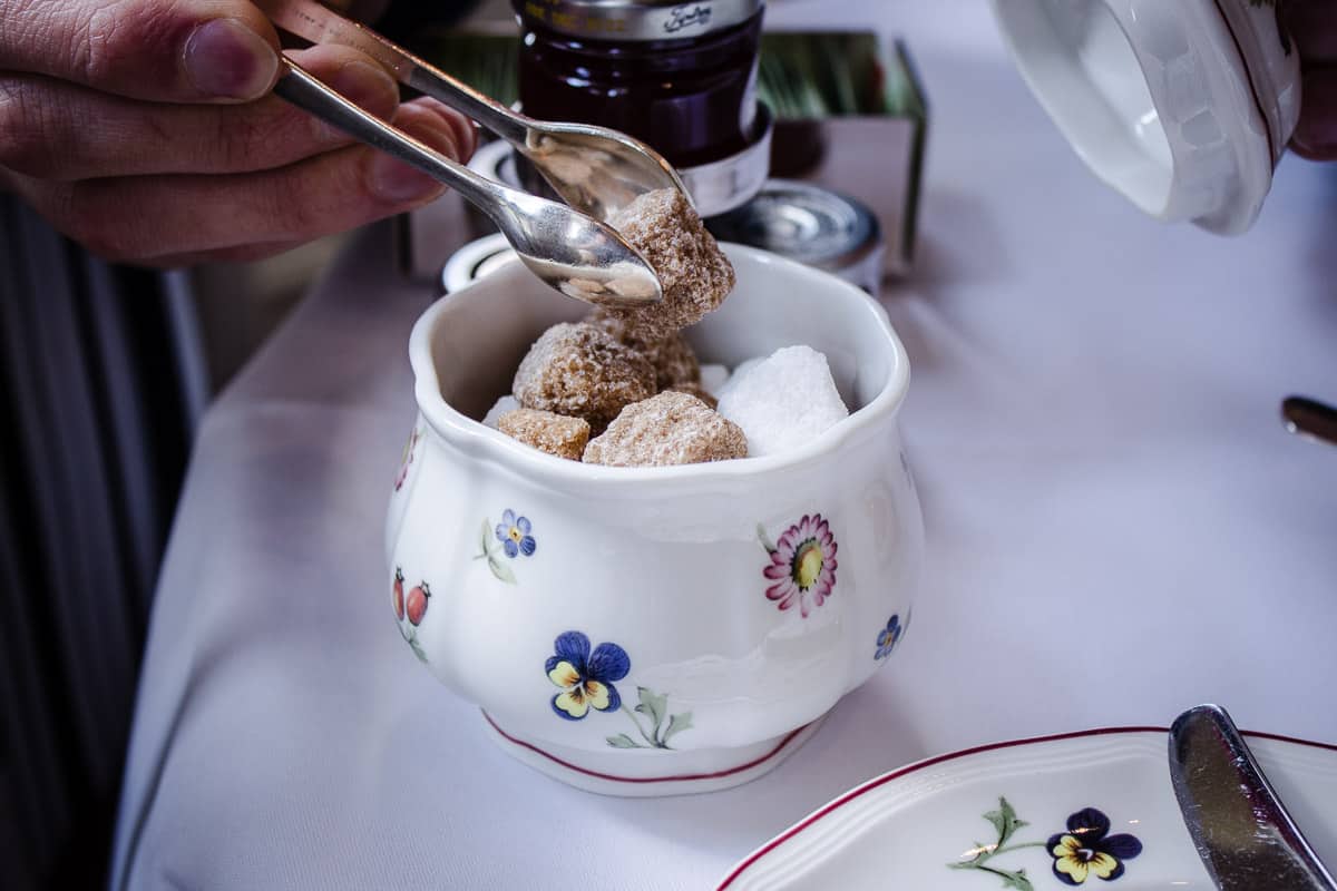 Image shows a bowl of sugar cubes from Egerton House's Vegan Afternoon Tea. A male hand is holding silver tongs and picking up a sugar cube.