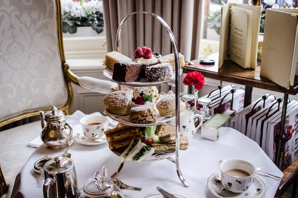 Image shows Egerton House's Vegan Afternoon Tea in London. There is a table set for two in a sitting room with a comfortable chair beside it. On the table is a three-tiered cake stand containing sandwiches, scones and cakes, surrounded by pots of tea and china cups/plates.