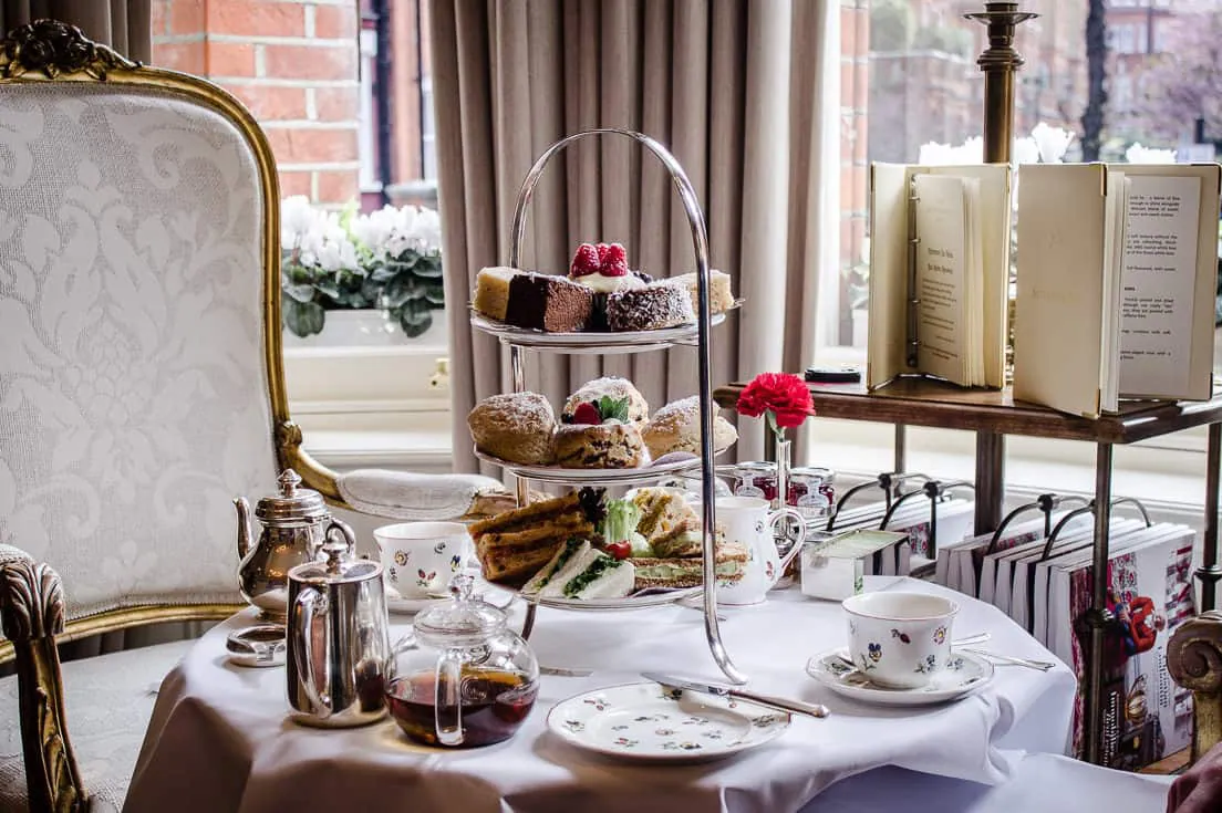 Image shows Egerton House's Vegan Afternoon Tea. There is a table set for two in a sitting room with a comfortable chair beside it. On the table is a three-tiered cake stand containing sandwiches, scones and cakes, surrounded by pots of tea and china cups/plates.