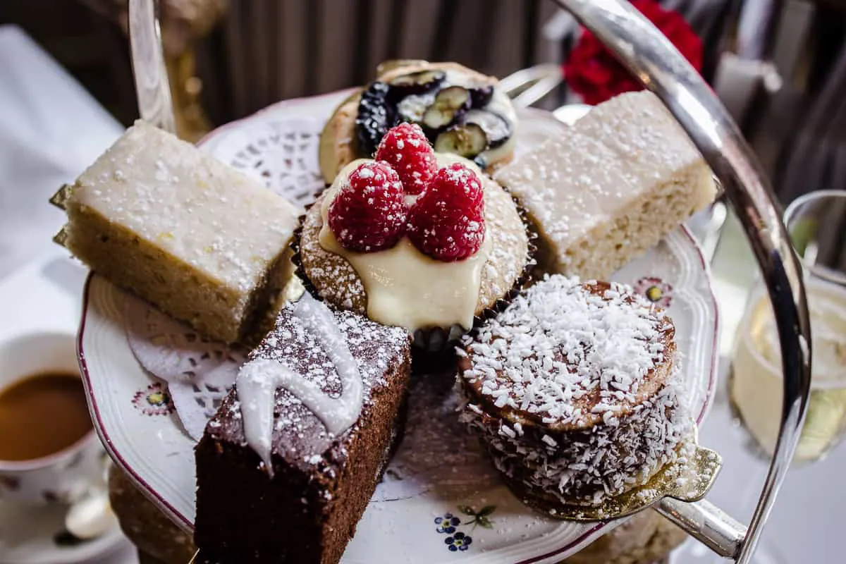Image shows a close-up of the cakes from Egerton House's Vegan Afternoon Tea. The top tier of a cake stand is shown, featuring a raspberry-topped cupcake, two rectangular lemon cakes, a blueberry tart and a chocolate brownie.
