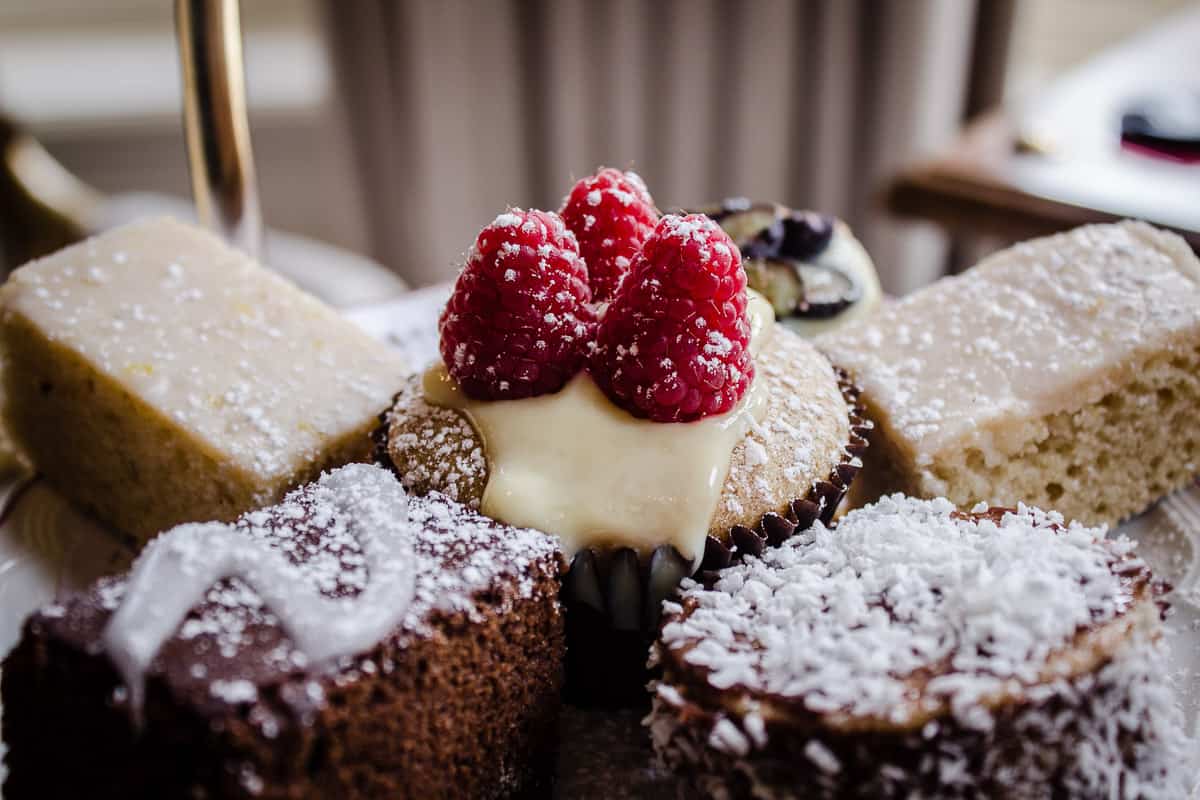Image shows a close-up of the cakes from Egerton House's Vegan Afternoon Tea in London. The top tier of a cake stand is shown, featuring a raspberry-topped cupcake, two rectangular lemon cakes, a blueberry tart and a chocolate brownie.
