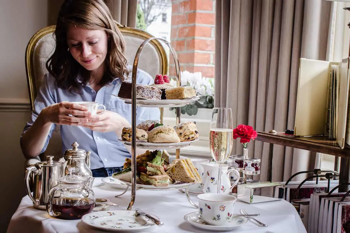 Image shows Egerton House's Vegan Afternoon Tea in London. There is a table set for two in a sitting room with a comfortable chair beside it. A woman is sitting in the chair clasping a cup of tea. On the table is a three-tiered cake stand containing sandwiches, scones and cakes, surrounded by a glass of champagne, pots of tea and china cups/plates.