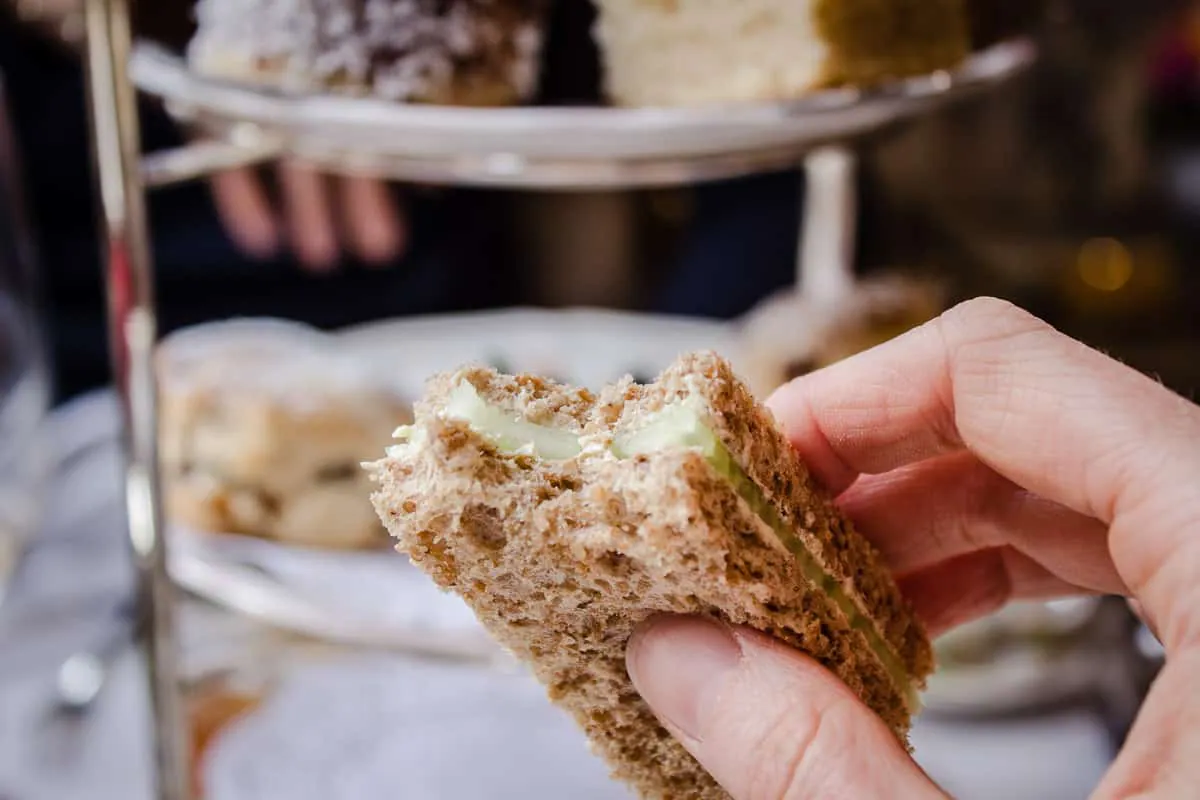 Image shows a finger sandwich from Egerton House's Vegan Afternoon Tea. The sandwich is filled with cucumber and is being held by a woman's hand. A cake stand is visible in the background.