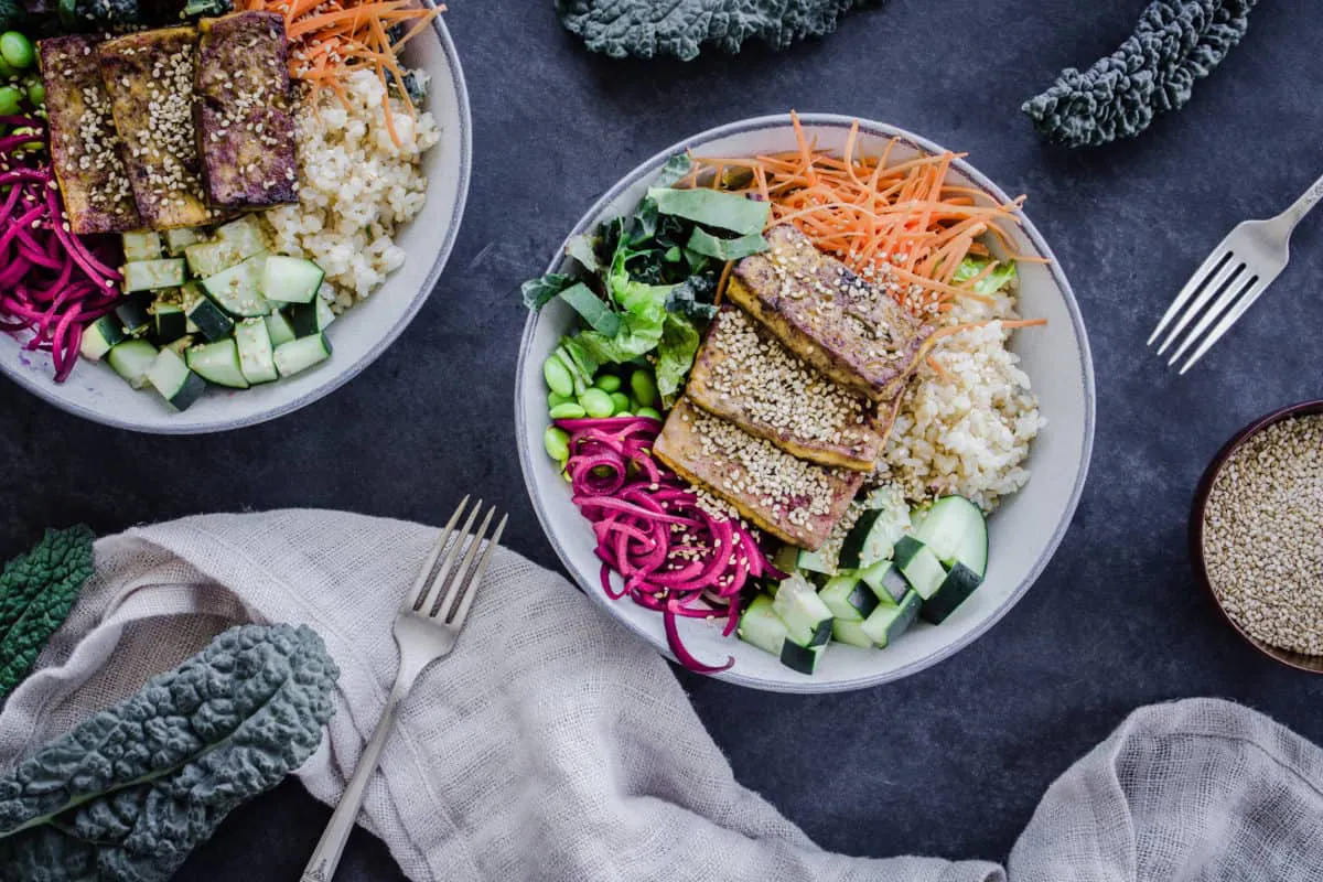 Two colourful vegan buddha bowls containing rice, vegetables and tofu, surrounded by forks, napkins, sesame seeds and kale leaves.
