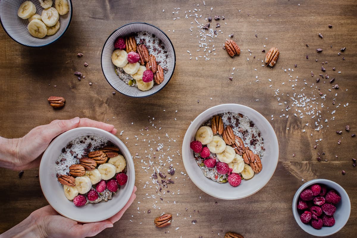 Overhead shot of bowls of Chocolatey Pecan Chia Cereal and woman's hands, decorated with fruit, coconut and nuts.