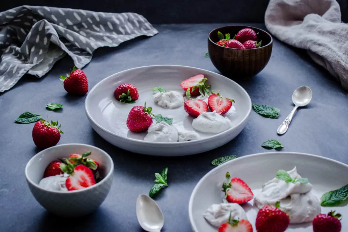 Plates and bowls of Easy Vegan Strawberries & Cream, surrounded by spoons, mint leaves, strawberries and napkins.