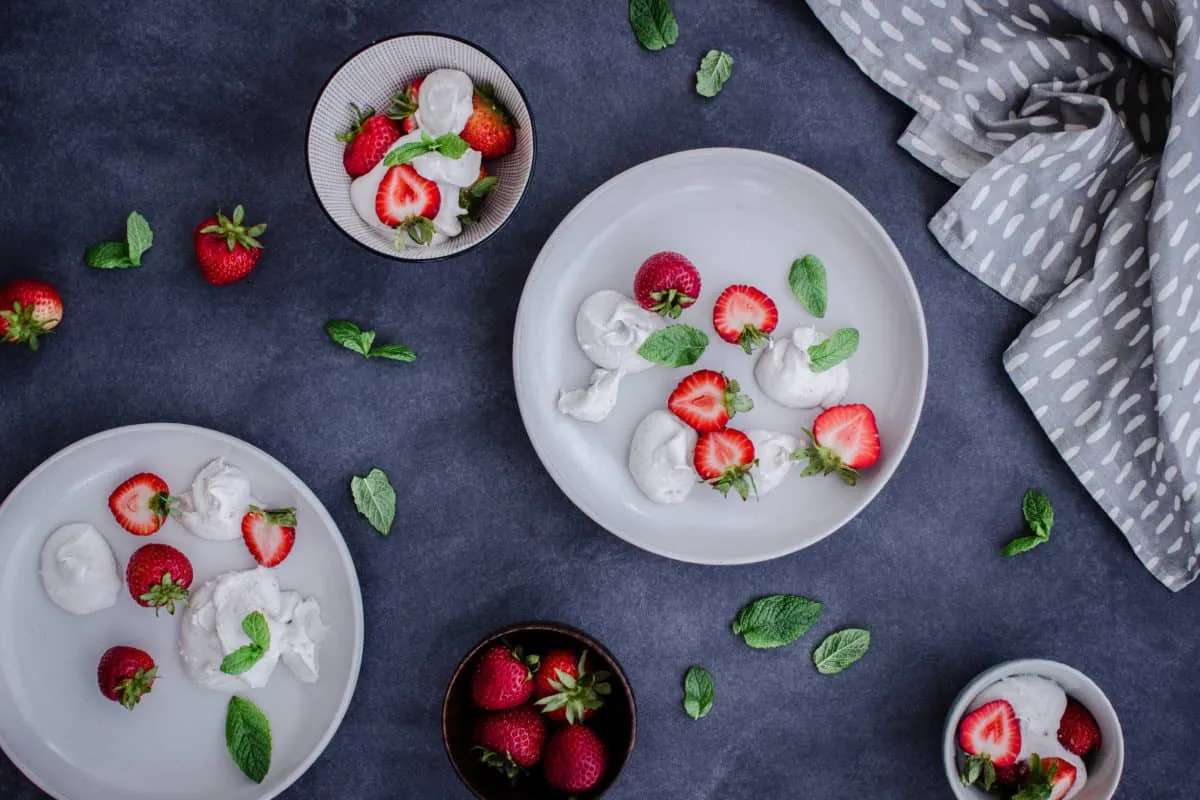 Overhead shot of plates and bowls of Easy Vegan Strawberries & Cream, surrounded by mint leaves, strawberries and napkins.