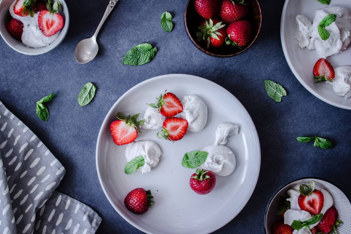 Overhead shot of plates and bowls of Easy Vegan Strawberries & Cream, surrounded by mint leaves, strawberries and napkins.