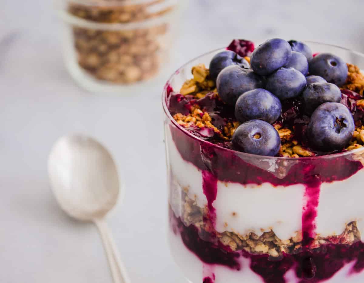 Close-up of Blueberry Breakfast Parfait consisting of layers of coconut yoghurt, blueberry compote, granola and fresh blueberries in a glass. Surrounded by a silver spoon and glass jar of granola.