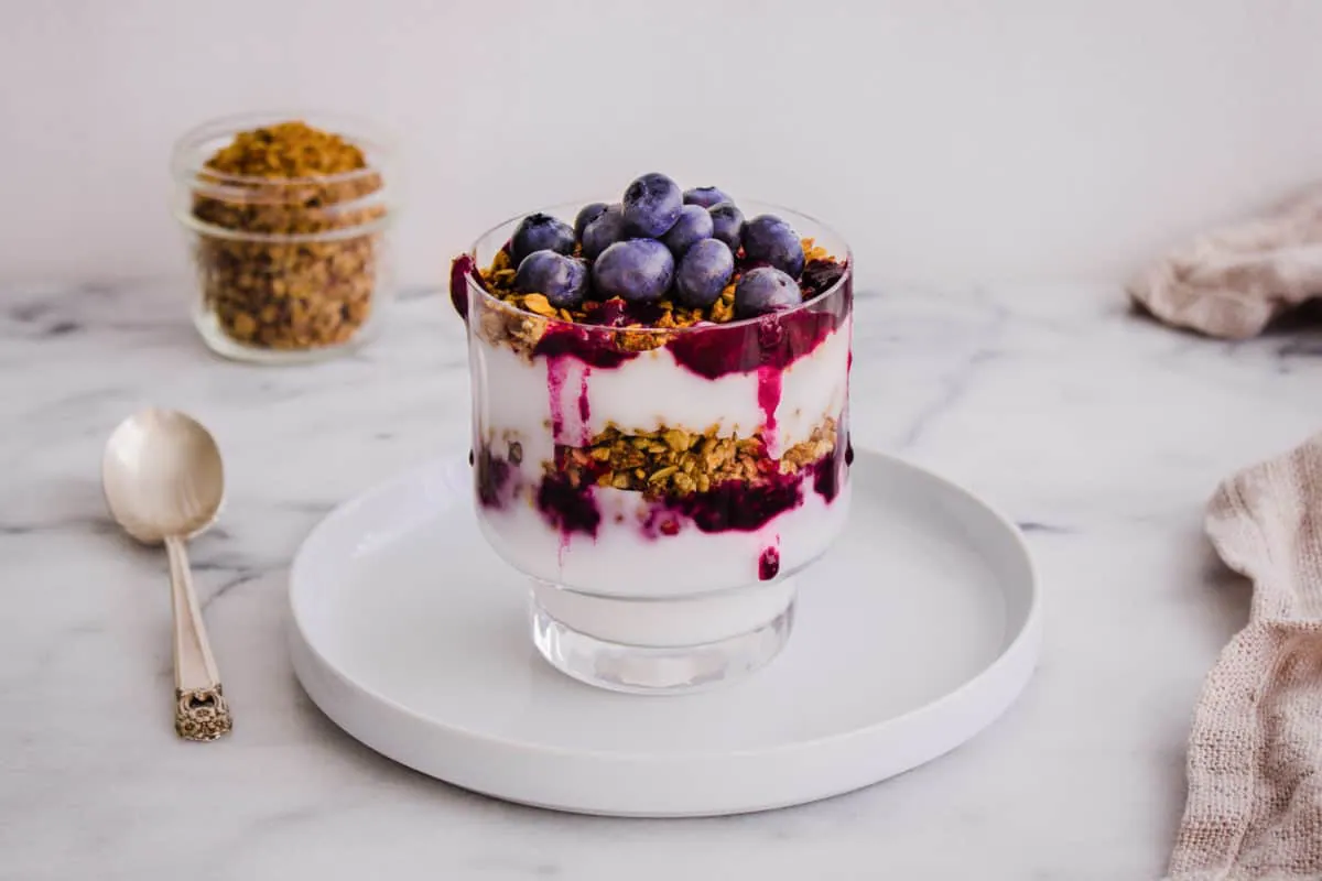 Image of Blueberry Breakfast Parfait consisting of layers of coconut yoghurt, blueberry compote, granola and fresh blueberries in a glass. Presented on a white plate, surrounded by a silver spoon, beige linen napkin and glass jar of granola..
