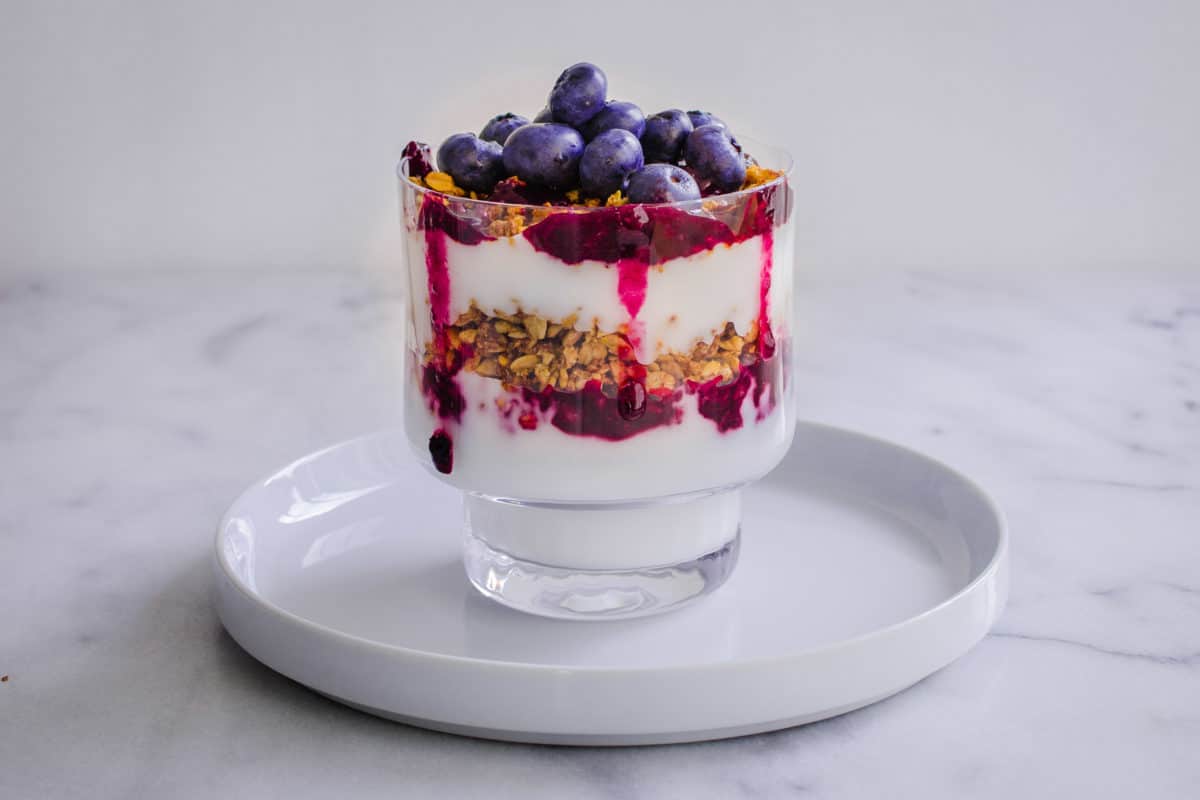 Image of Blueberry Breakfast Parfait consisting of layers of coconut yoghurt, blueberry compote, granola and fresh blueberries in a glass. Presented on a white plate, with white marble surface in the background.