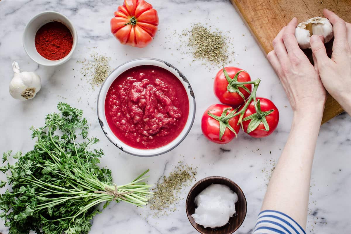 How to Go Vegan (10 Easy Steps). Image shows ingredients to make homemade tomato pasta sauce on a white marble surface. A woman's hand can be seen holding a bulb of garlic on a chopping board.