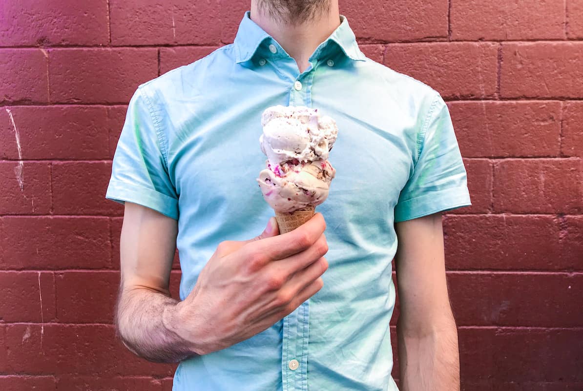 Man in a bright blue shirt against a red brick wall holds an ice cream cone. The ice cream has two scoops: one is white, one is pink studded with nuts.