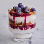 Image of Blueberry Breakfast Parfait consisting of layers of coconut yoghurt, blueberry compote, granola and fresh blueberries in a glass, with white marble surface in the background.