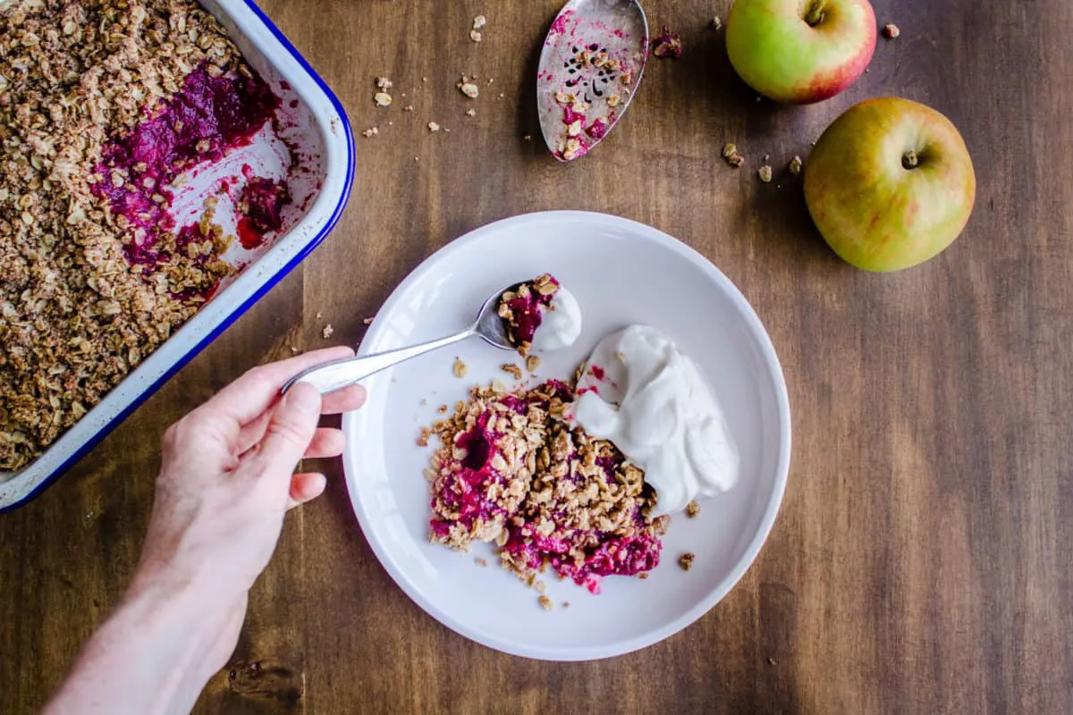 Overhead shot of Apple & Blackberry Crumble. Image shows a woman's hand holding a spoon of crumble which has a pink apple and blackberry filling. Below the spoons there is a white plate with a serving of crumble and coconut yoghurt. The dish is surrounded by a white enamel baking tray containing the rest of the crumble, an old silver cake slice and some apples, and everything is sitting on a rustic wooden table.