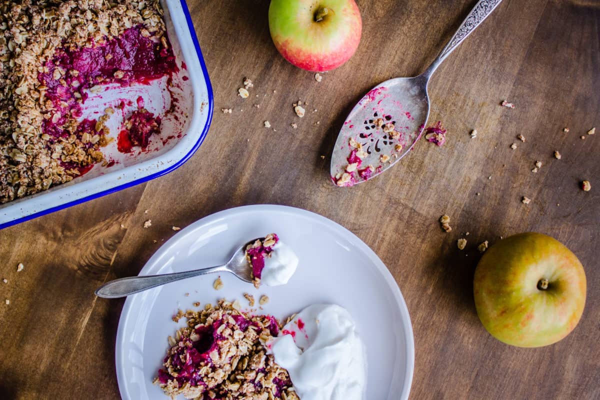 Overhead image of Apple & Blackberry Crumble. Image shows the edge of a tray of crumble with a pink filling, and next to it a white plate with a serving of crumble and coconut yoghurt. The dishes are surrounded by old silver cutlery and apples, and are sitting on a wooden table.