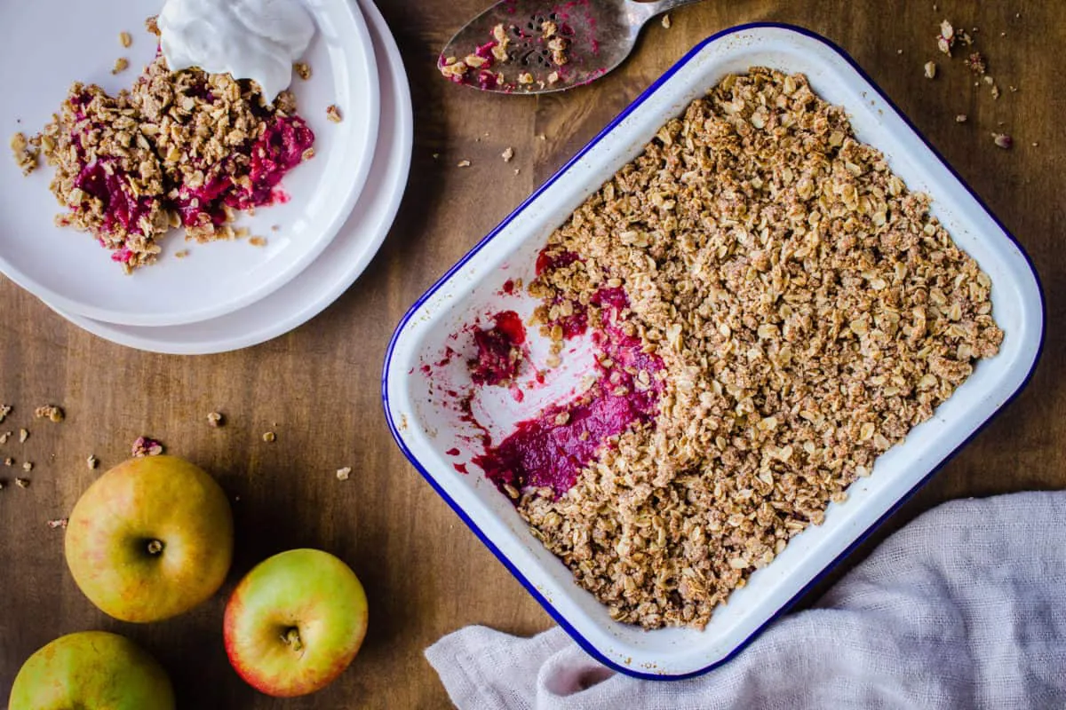 Overhead image of Apple & Blackberry Crumble. Image shows a white enamel tray of crumble with a pink apple and blackberry filling, and next to it a white plate with a serving of crumble and coconut yoghurt. The dishes are surrounded by an old silver cake slice, linen napkin, and apples, and are sitting on a wooden table.
