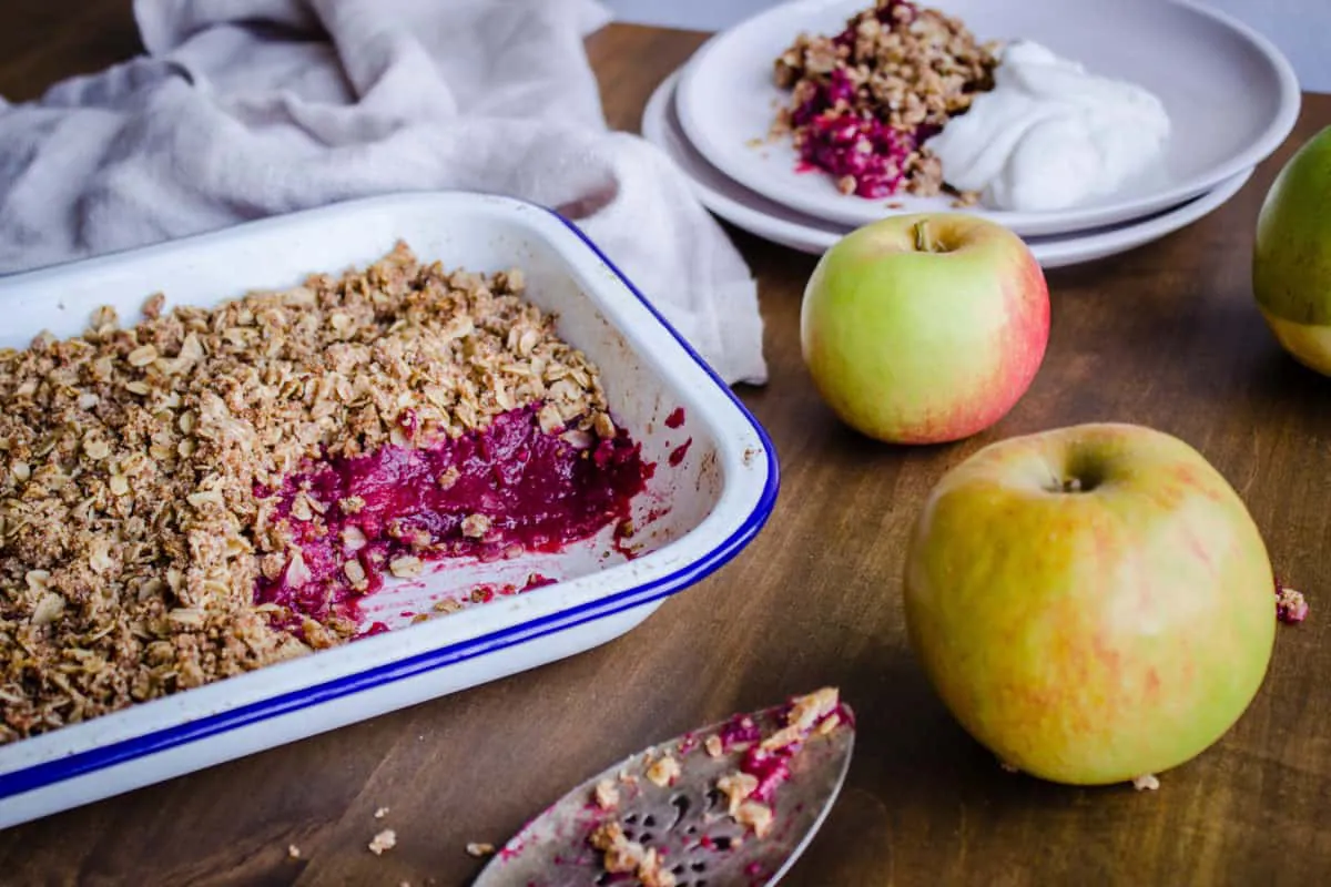 Closeup shot of Apple & Blackberry Crumble. Image shows a white enamel tray of crumble with a pink apple and blackberry filling, surrounded by white plates with a serving of crumble and coconut yoghurt. The dishes are surrounded by an old silver cake slice, a linen napkin, and some apples, and are sitting on a rustic wooden table.