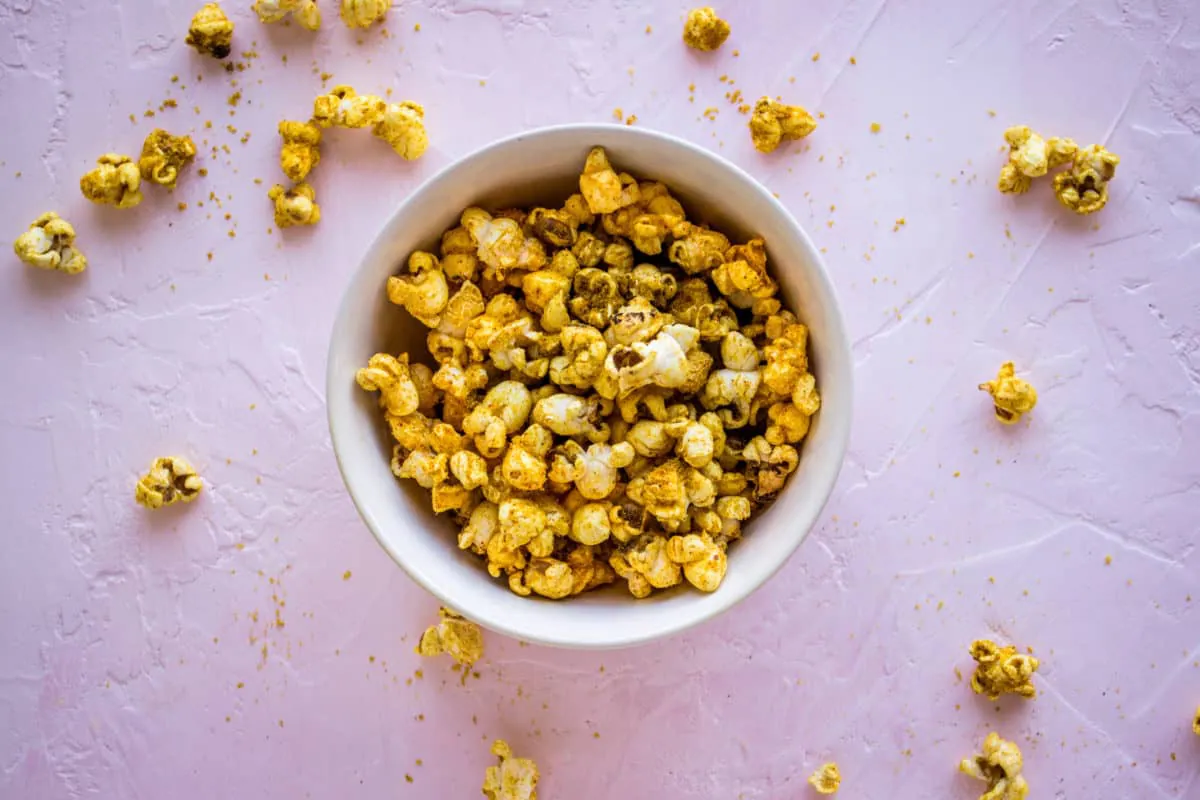 Overhead shot of white bowl of Cheesy Vegan Popcorn on light pink plaster background, surrounded by scattered pieces of popcorn.