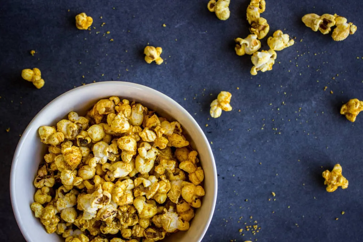 Overhead shot of white bowl of Cheesy Vegan Popcorn on black slate background, surrounded by scattered pieces of popcorn.