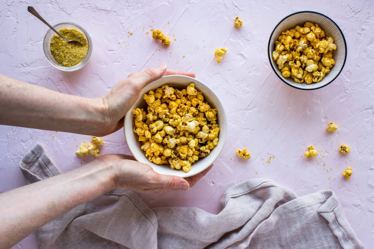 Overhead shot of two white bowls of Cheesy Vegan Popcorn with a woman's hands clasping one, on a light pink plaster background, surrounded by scattered pieces of popcorn, a linen napkin and a glass jar of nutritional yeast.