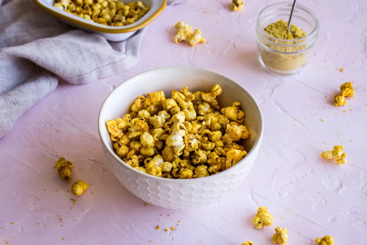 Image of two white bowls of Cheesy Vegan Popcorn on light pink plaster background, surrounded by scattered pieces of popcorn, a linen napkin and a glass jar of nutritional yeast.