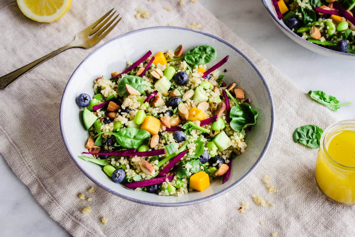 Close-up image of Quinoa Power Bowls, showing two grey bowls with quinoa, bright-coloured vegetables, fruit and toasted nuts in them, on a beige linen napkin accompanied by spinach leaves, quinoa, a lemon wedge and bright yellow vinaigrette.