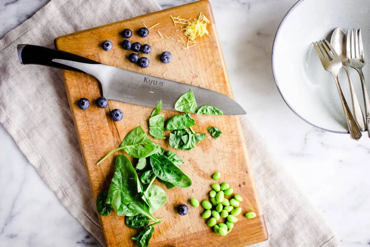 Overhead image showing the prep for Quinoa Power Bowls, showing a wooden chopping board and sharp knife with spinach, blueberries, edamame and lemon zest scattered across it. The chopping board is on a beige linen napkin, and is accompanied by a light grey bowl and silver cutlery on a white marble background.