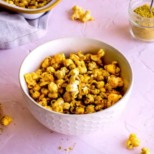 White bowl of Cheesy Vegan Popcorn on light pink plaster background, surrounded by scattered pieces of popcorn, a linen napkin and a glass jar of nutritional yeast.