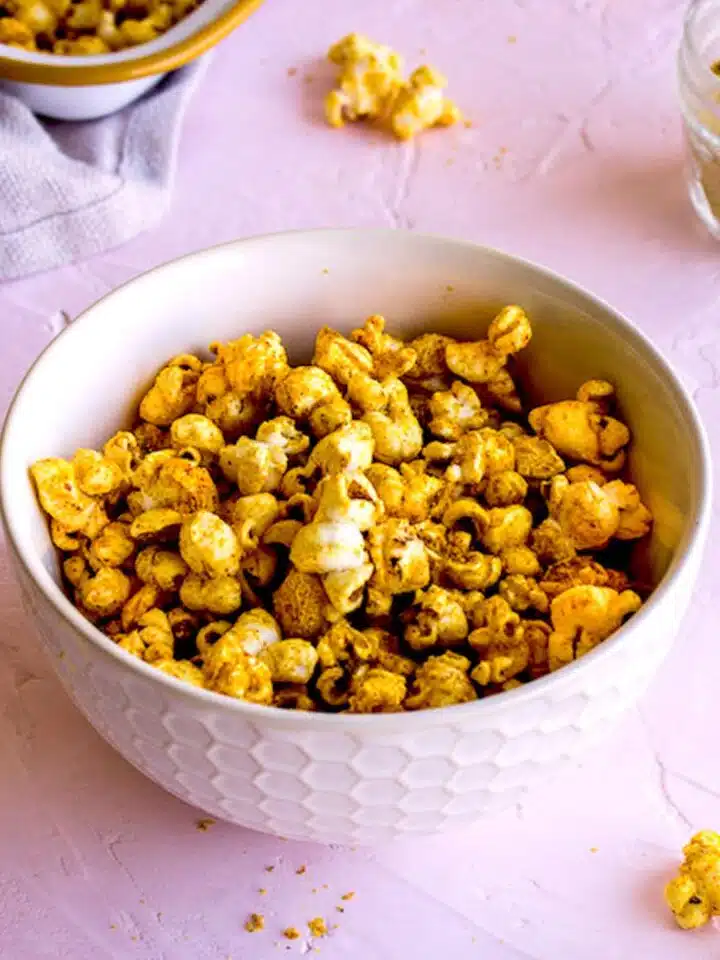 White bowl of Cheesy Vegan Popcorn on light pink plaster background, surrounded by scattered pieces of popcorn, a linen napkin and a glass jar of nutritional yeast.