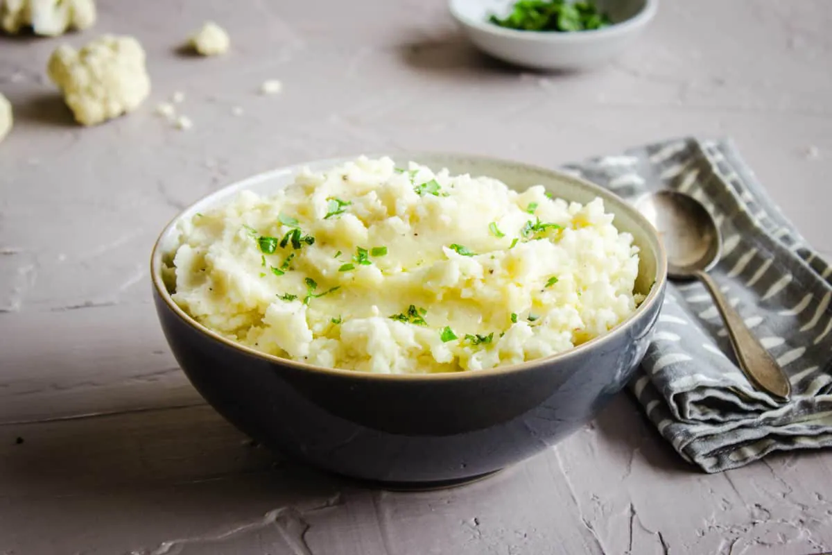 Image shows a grey bowl of Cauliflower Mashed Potatoes sprinkled with parsley and drizzled with olive oil. The bowl is on a grey plaster background and is surrounded by a bowls of parsley, a napkin, a silver spoon and cauliflower florets.