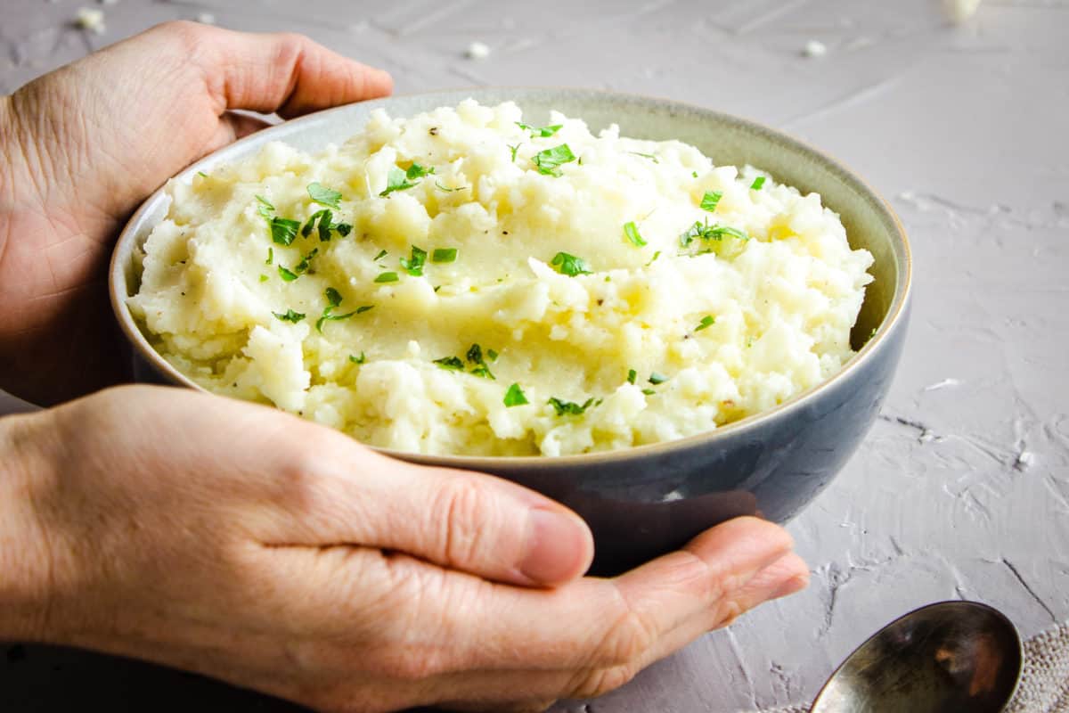 Image shows a bowl of Cauliflower Mashed Potatoes sprinkled with parsley and drizzled with olive oil. A woman's hands are holding the bowl and it is on a grey plaster background.