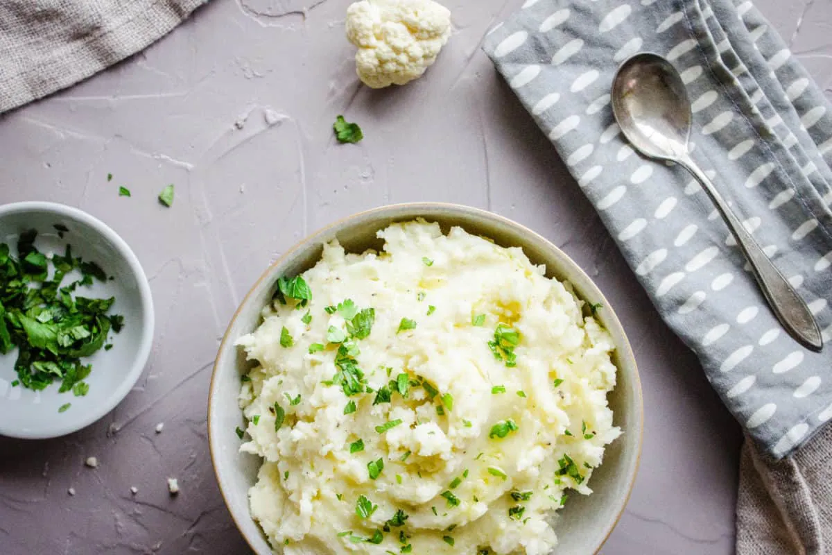Overhead image shows a bowl of Cauliflower Mashed Potatoes sprinkled with parsley and drizzled with olive oil. The bowl is on a grey plaster background and is surrounded by a bowls of parsley, napkins, a silver spoon and cauliflower florets.