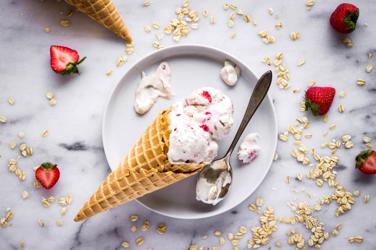 Overhead image of Vegan Strawberry Ice Cream with Cookie Dough. Image shows a waffle cone lying on a white plate, on a white marble surface with two scoops of ice cream in it. The cone is surrounded by an antique silver spoon and drips of ice cream. Also visible in the image is another cone, some strawberries and some oats.