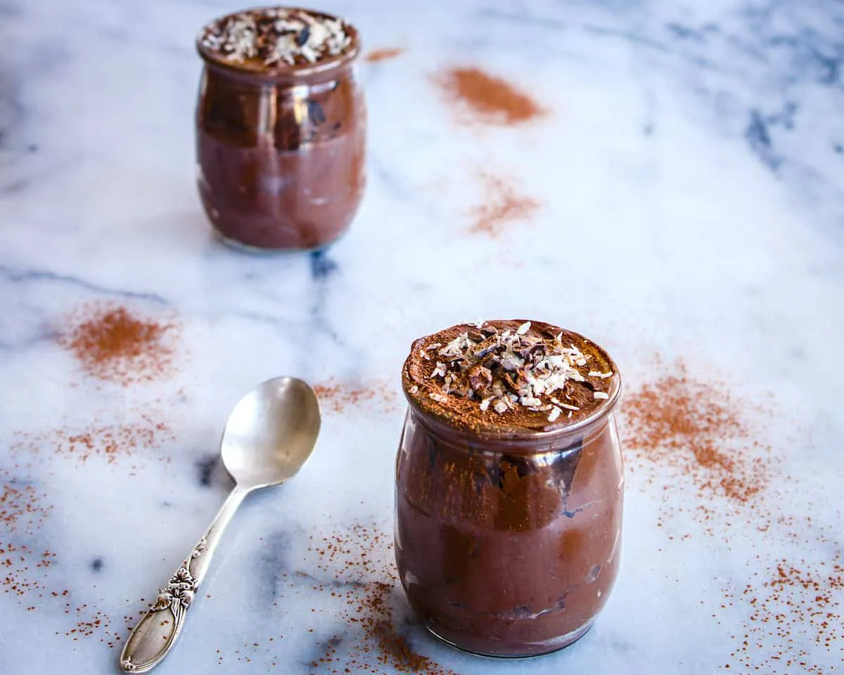 Image of Avocado Chocolate Mousse (vegan & gf). Image shows two pots of chocolate mousse on a white marble table, surrounded by piles of cacao and an antique silver spoon.