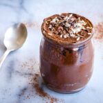 Image of Avocado Chocolate Mousse (vegan & gf). Image shows one pot of chocolate mousse on a white marble table, surrounded by piles of cacao powder and an antique silver spoon.