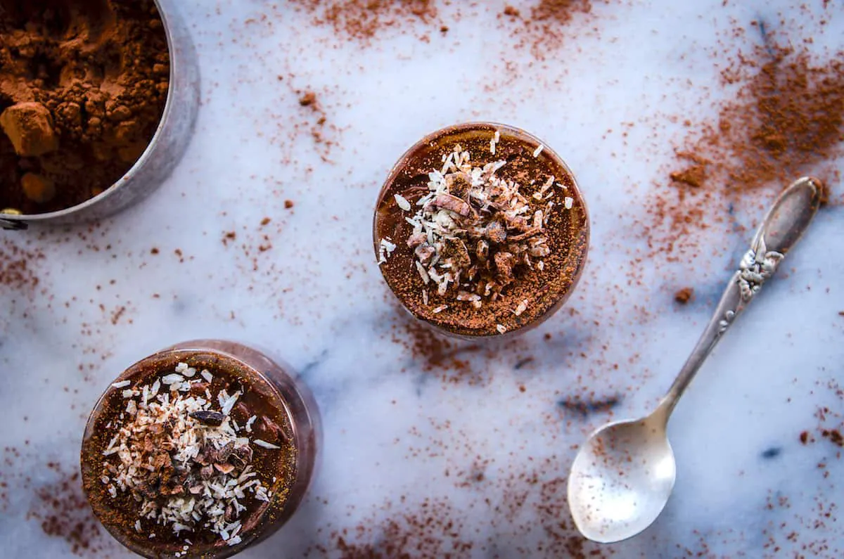 Overhead image of Avocado Chocolate Mousse (vegan & gf). Image shows two pots of chocolate mousse on a white marble table, surrounded by a cup of cacao powder, piles of cacao and an antique silver spoon.