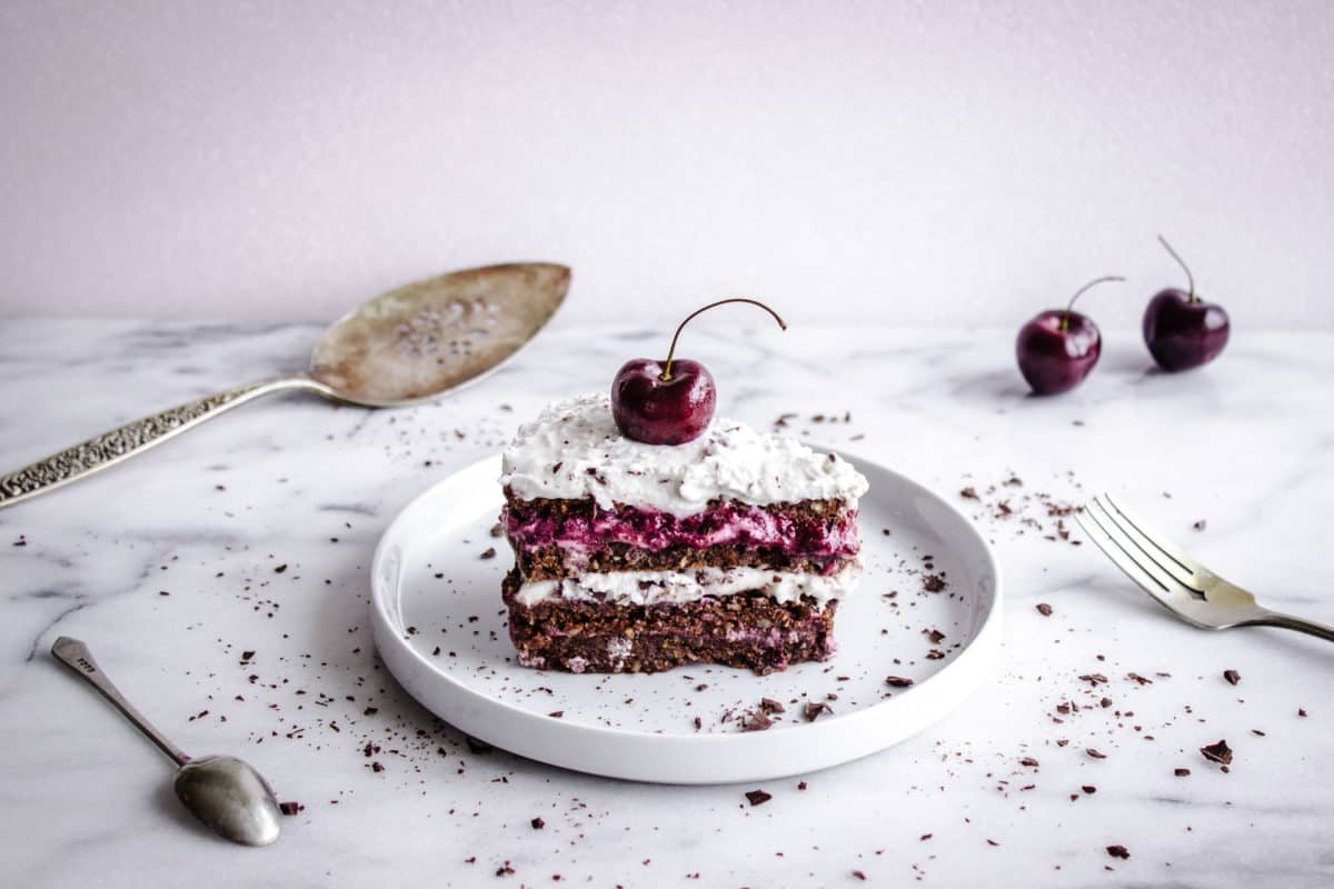 Shot of No-Bake Black Forest Cake, showing slice of chocolate-flavoured raw cake with layers of cherry jam and coconut cream. The cake is decorated with a whole cherry and chocolate shavings. It is placed on a white plate on a white marble countertop, and is surrounded by silver cultery, a cake slice, cherries and more chocolate shavings.