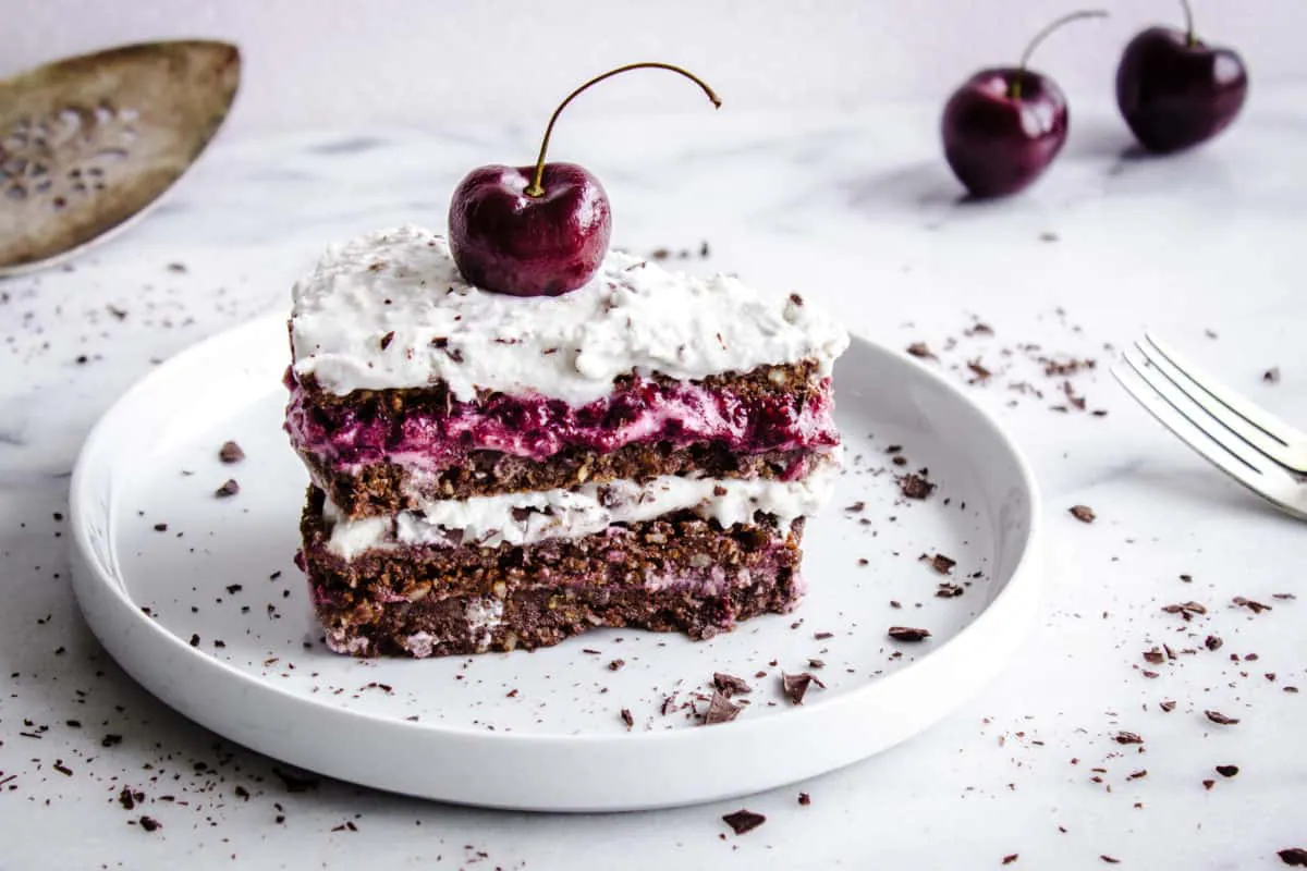 Close-up shot of No-Bake Black Forest Cake, showing slice of chocolate-flavoured raw cake with layers of cherry jam and coconut cream. The cake is decorated with a whole cherry and chocolate shavings. It is placed on a white plate on a white marble countertop, and is surrounded by silver cutlery, a cake slice, cherries and more chocolate shavings.