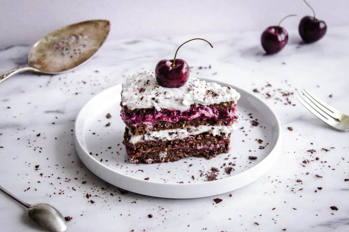 Shot of No-Bake Black Forest Cake, showing slice of chocolate-flavoured raw cake with layers of cherry jam and coconut cream. The cake is decorated with a whole cherry and chocolate shavings. It is placed on a white plate on a white marble countertop, and is surrounded by silver cutlery, a cake slice, cherries and more chocolate shavings.
