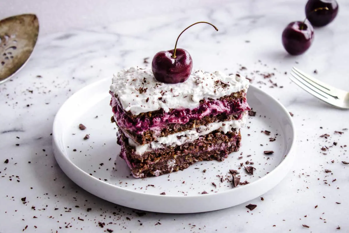 Shot of No-Bake Black Forest Cake, showing slice of chocolate-flavoured raw cake with layers of cherry jam and coconut cream. The cake is decorated with a whole cherry and chocolate shavings. It is placed on a white plate on a white marble countertop, and is surrounded by silver cutlery, a cake slice, cherries and more chocolate shavings.