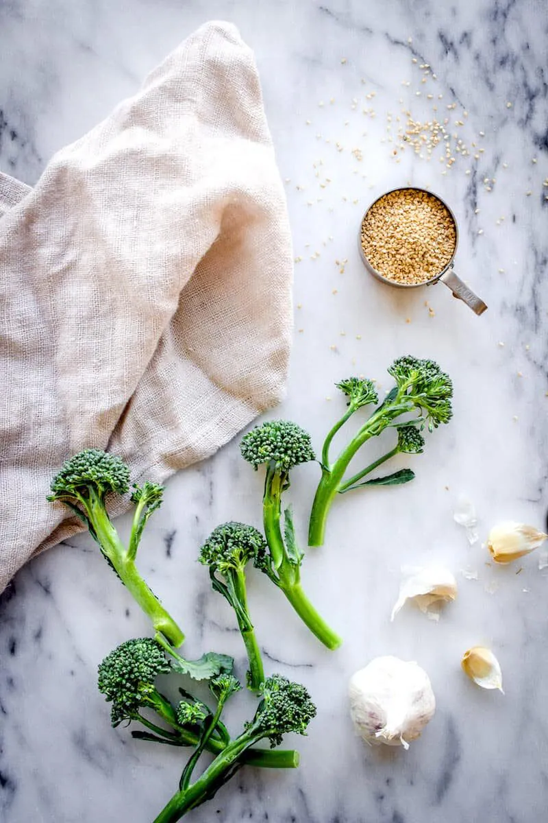 Overhead image of ingredients to make Roasted Broccoli with Sesame & Garlic. Stalks of broccolini, cloves of garlic and a cup of golden sesame seeds are visible on a white marble background. Next to them is a linen napkin.