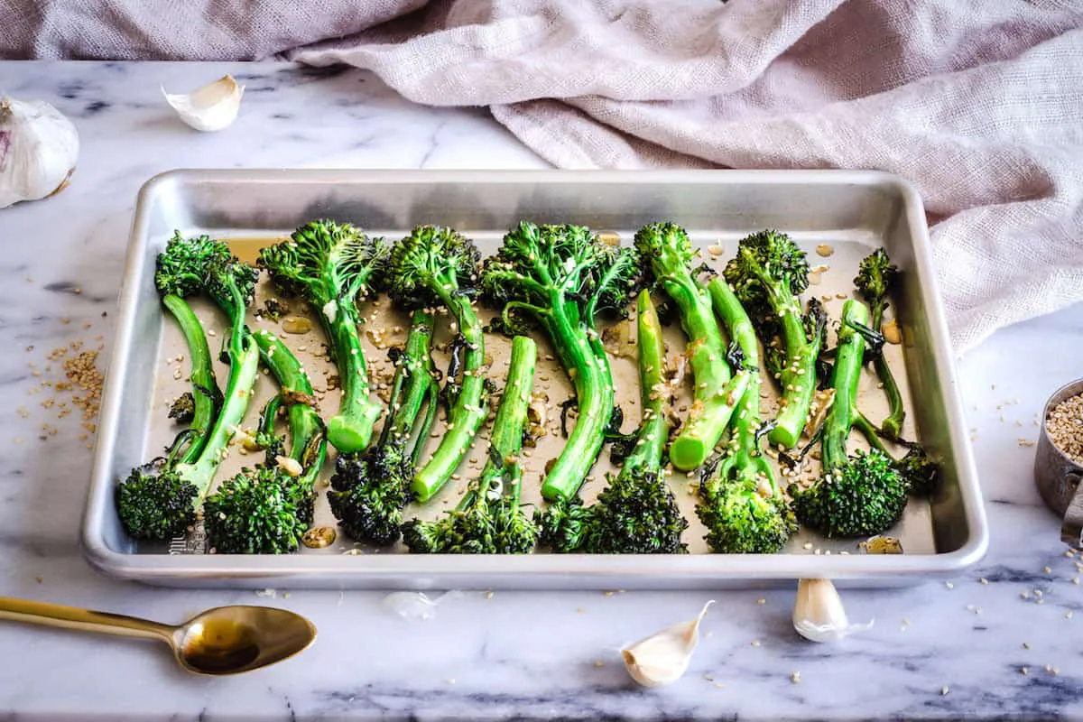Image of Roasted Broccoli with Sesame & Garlic. A silver baking tray of roasted broccoli is visible on a white marble background. Surrounding it is a golden spoon, a cup of sesame seeds, some garlic cloves and a linen napkin.