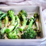 Closeup image of Roasted Broccoli with Sesame & Garlic. A silver baking tray of roasted broccoli is visible on a white marble background. Surrounding it are some sesame seeds and a linen napkin.